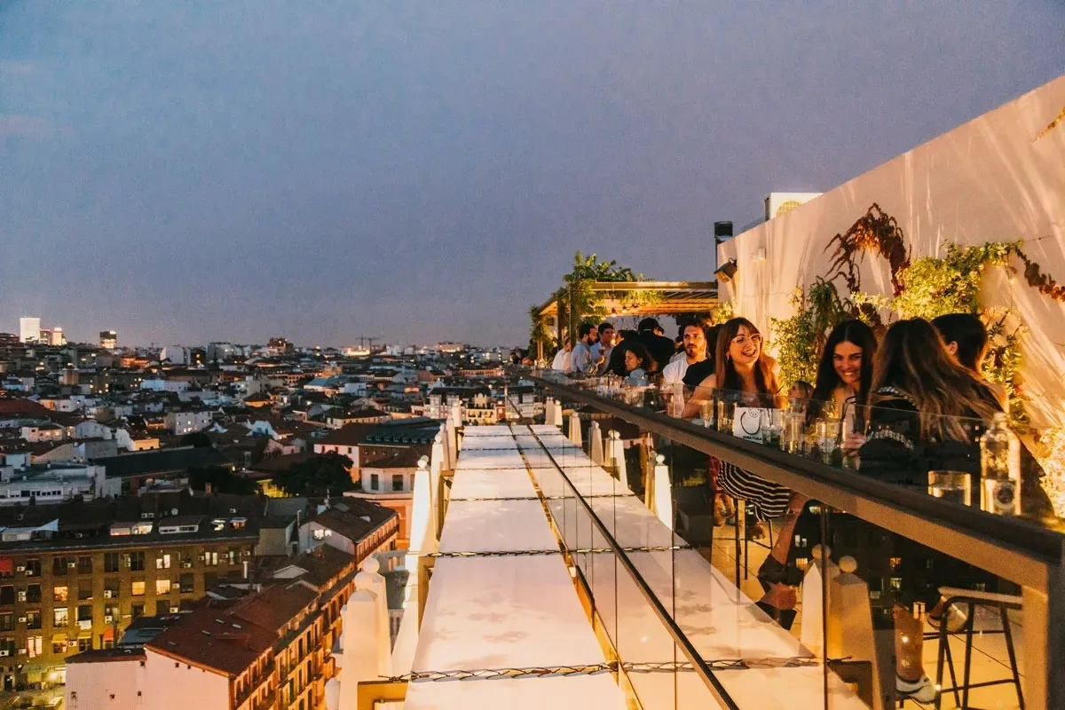 A long rooftop terrace with a bar and drinkers at dusk above the city lights