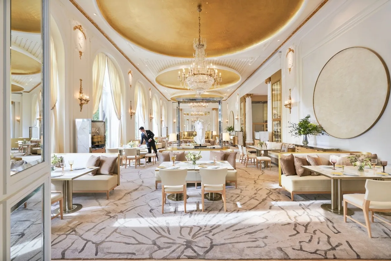 A long, bright dining room with a gilded domed ceiling, tall windows and tables set along the length of the room