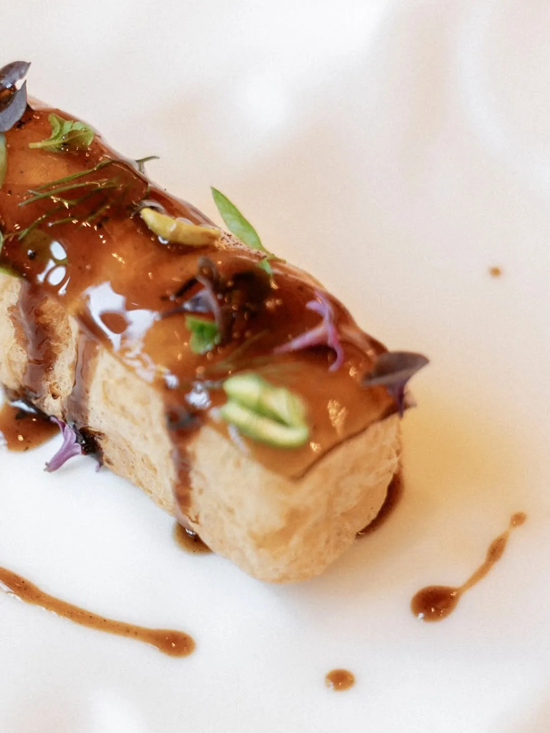 A glazed rectangular piece of food with green garnish and drops of sauce on a white plate