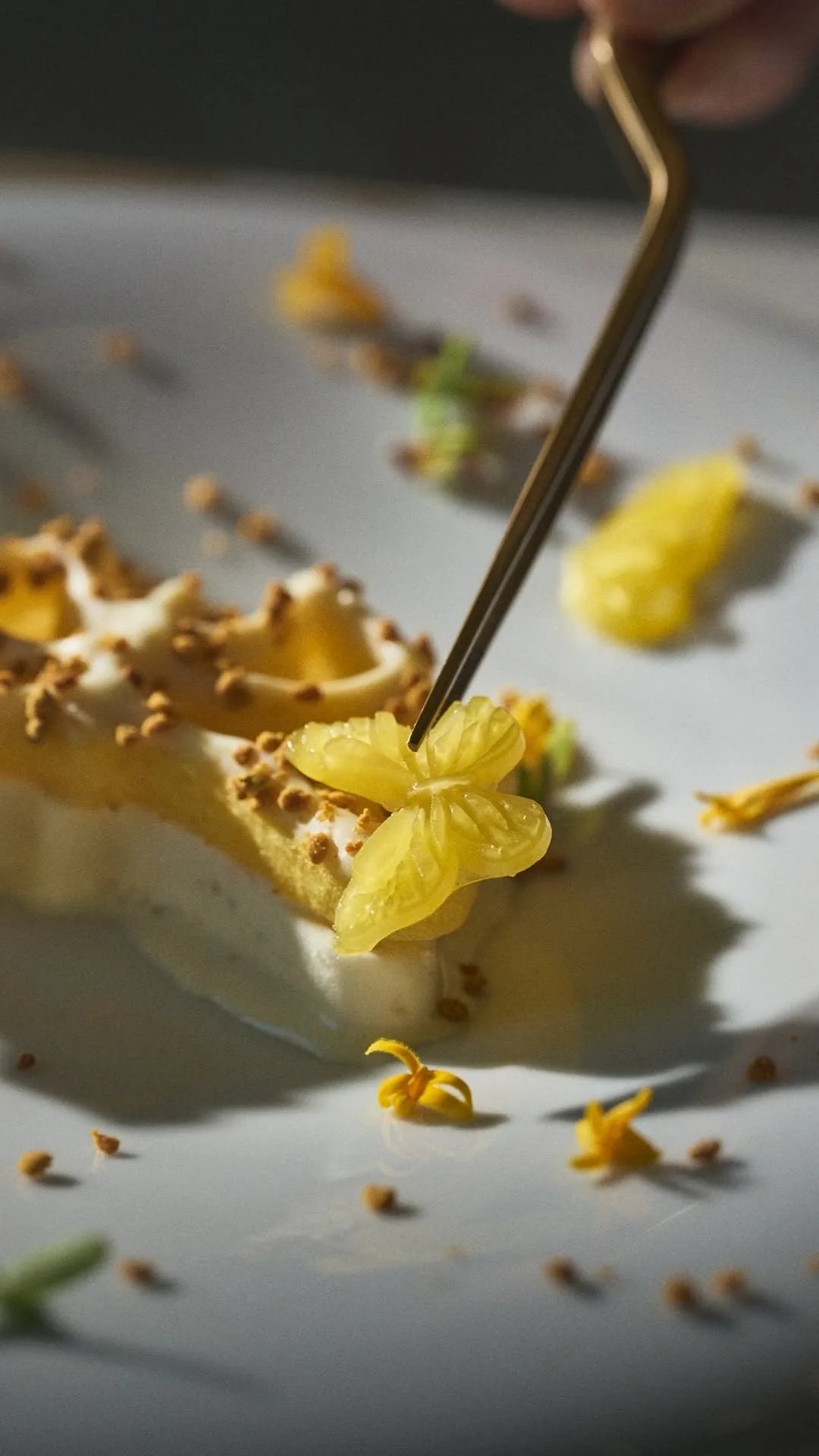 A dessert dish with golden flower petals and small sauce dots being finished with tweezers