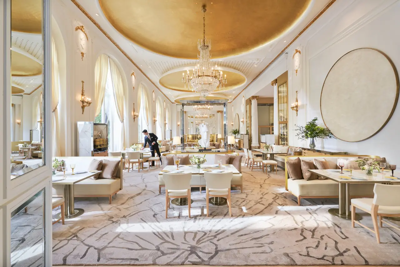 White-and-gold dining room under a gilded domed ceiling, with crystal chandeliers, tall arched windows, cream banquettes and set tables