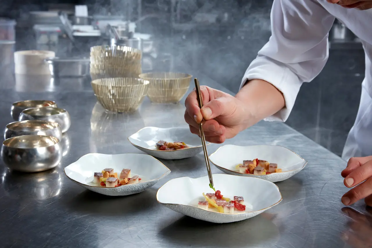 A chef plating small portions with tweezers into white ceramic bowls on a steel pass