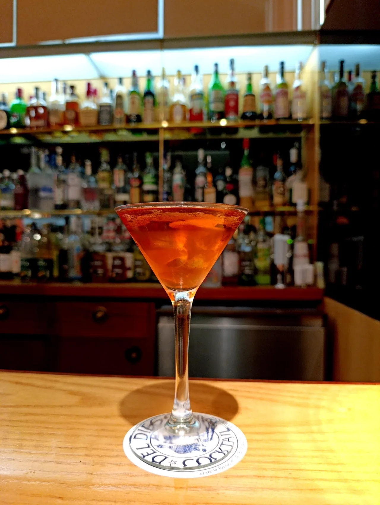 A red cocktail in a stemmed glass on a wooden bar with shelves of bottles behind