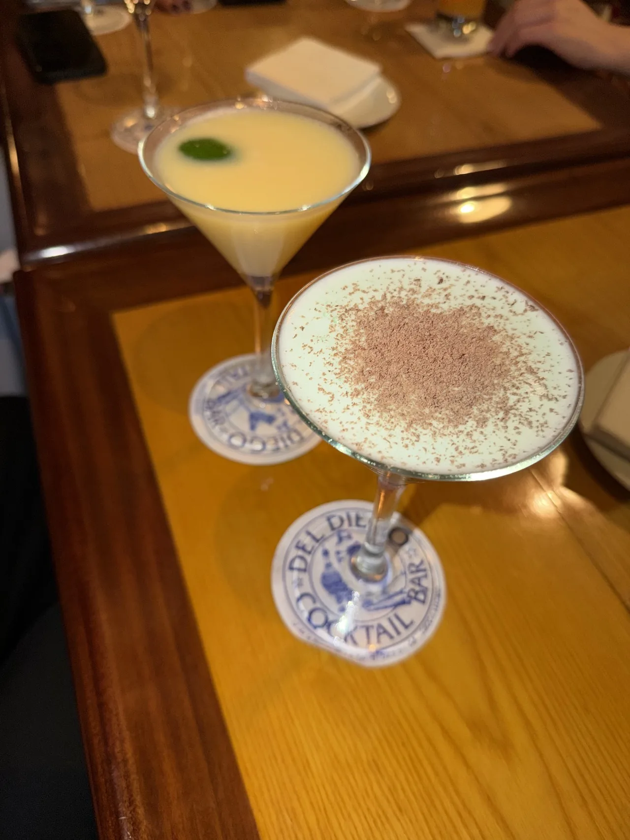 Two cocktails in coupe glasses on coasters on a wooden table
