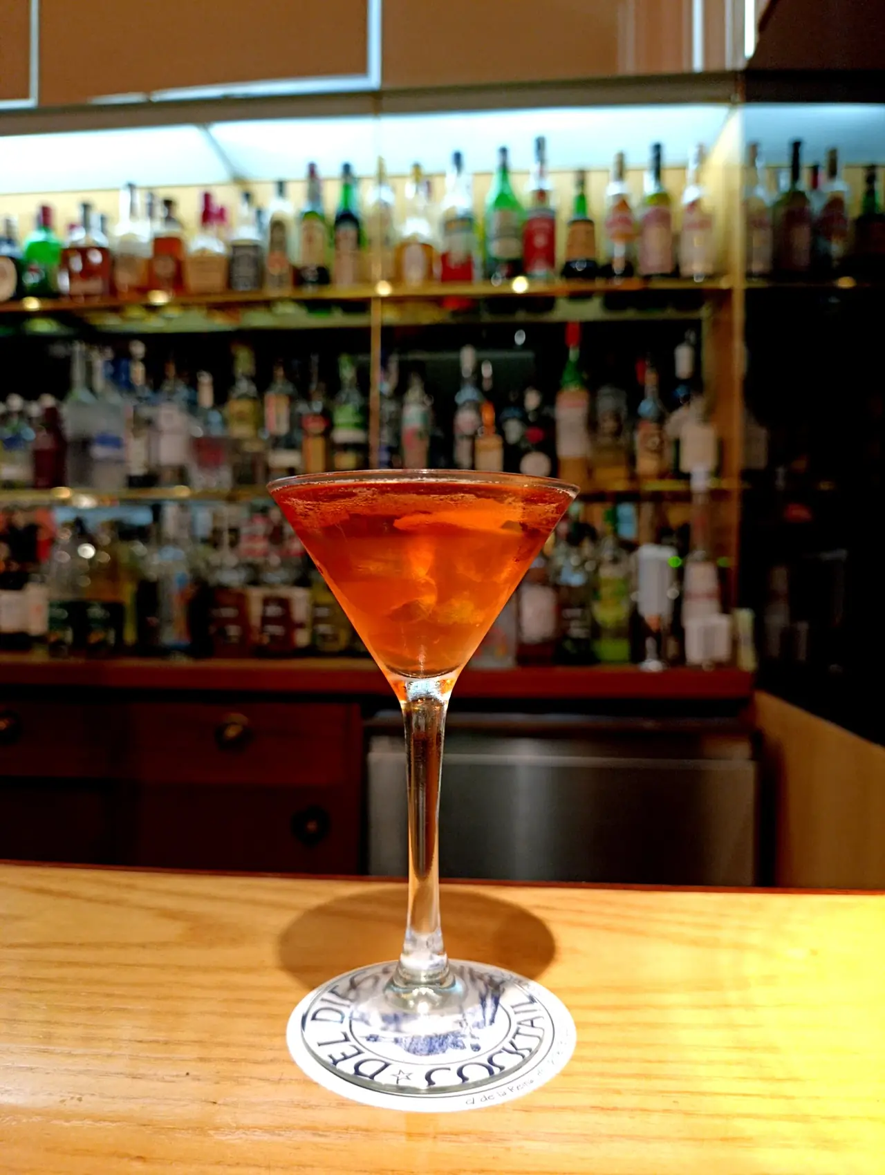 Amber cocktail in a martini glass on the bar, backed by shelves of spirits