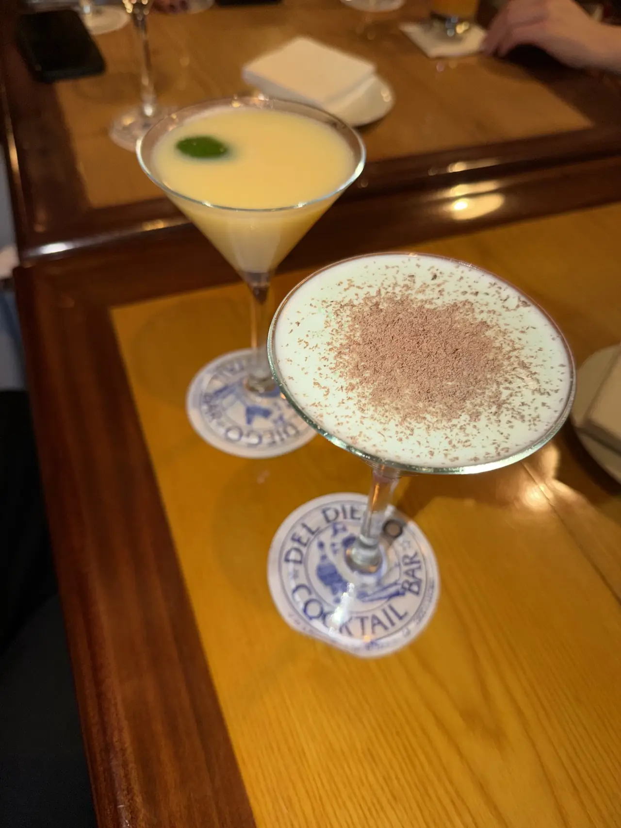 Two cocktails on Del Diego Cocktail Bar coasters, one pale yellow with a mint leaf, one creamy white dusted with cocoa powder