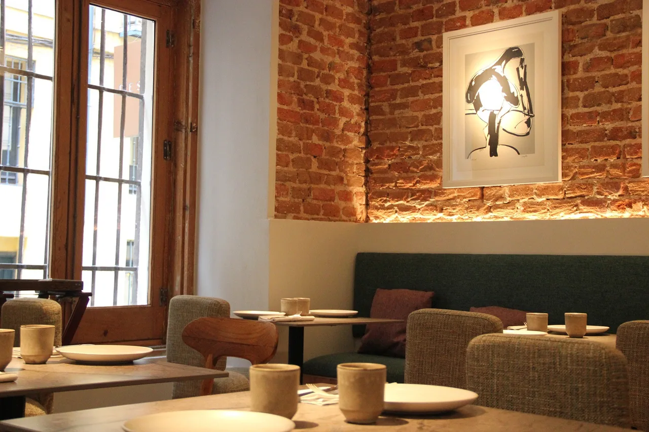 A corner of a dining room with a green banquette, wooden tables, a red brick wall and framed art