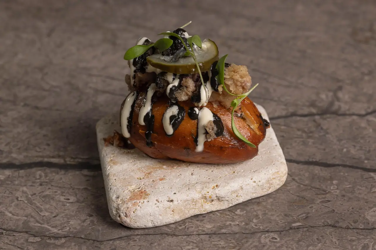 Glazed bun bite topped with crispy bits, sauce drizzle and a pickle slice on a stone board