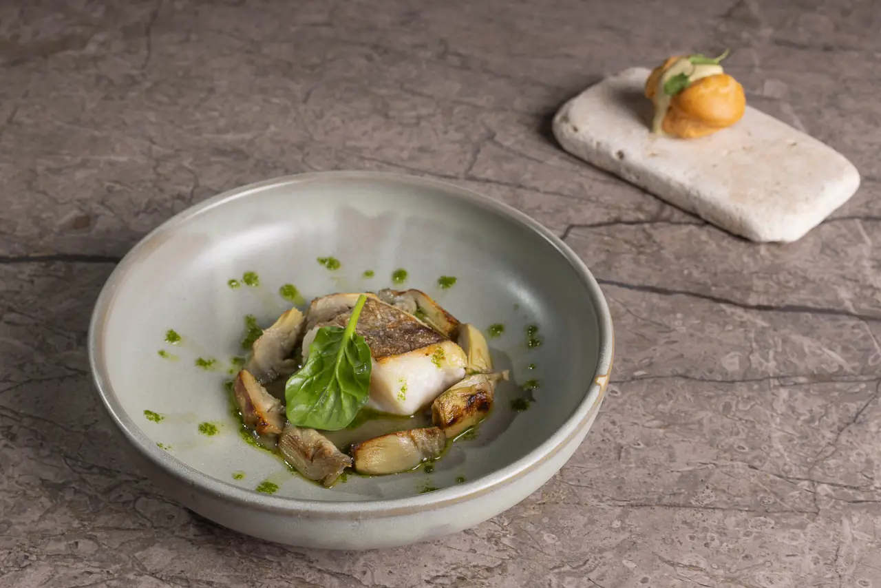 Seared fish fillet with artichoke pieces and green herb oil dots on a pale bowl