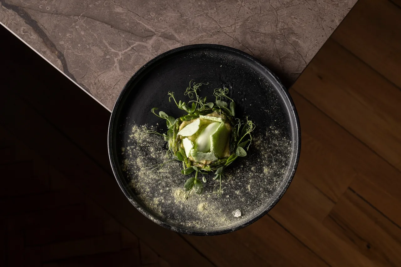 A top-down view of a black plate with asparagus, pea shoots, a green purée and pale green crisps