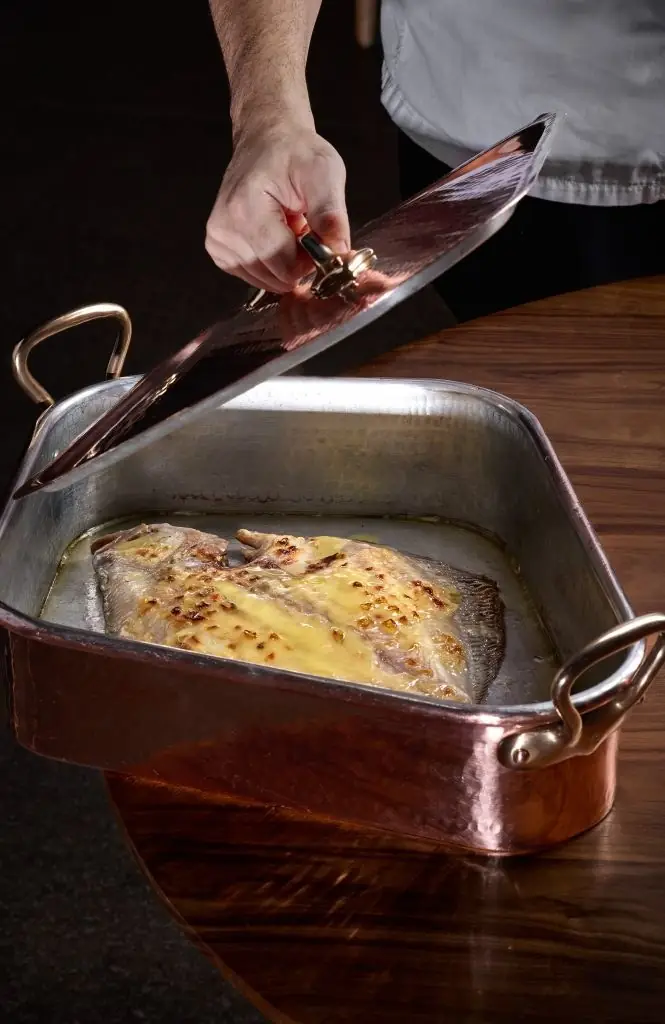 A whole flat fish glazed golden and bubbling in butter inside a copper and steel roasting pan, a cook lifting the hammered lid away above it