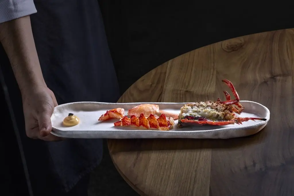A sliced lobster tail and a grilled half lobster on a long white ceramic platter with a quenelle of yellow sauce topped with caviar, carried by a waiter