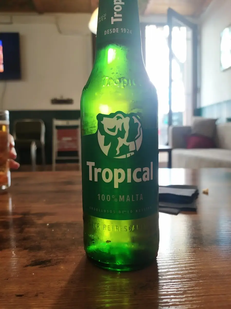 Green bottle of Tropical beer standing on a wooden table with the venue's interior and seating behind