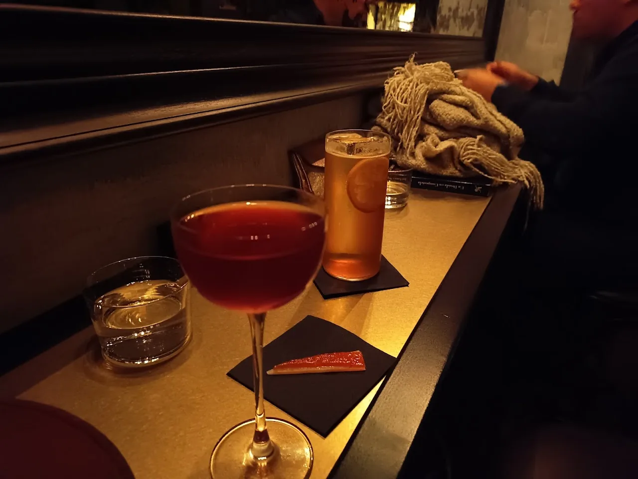 A reddish-brown cocktail on the bar counter with a beer in the background