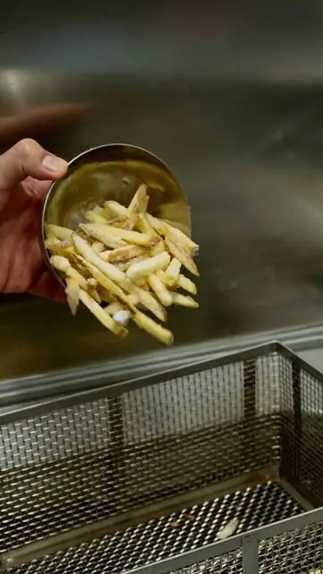 A hand tipping a metal scoop of French fries over a fryer basket in a stainless steel kitchen