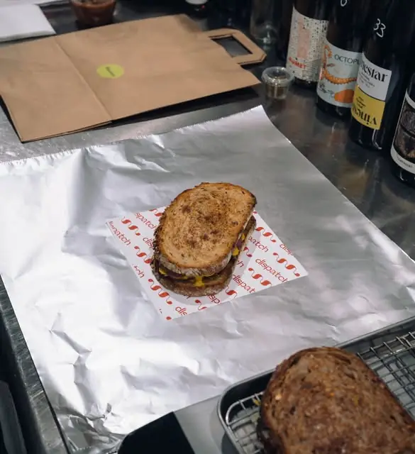 A toasted sandwich on Dispatch-branded paper and foil, with wine bottles and a takeaway bag behind