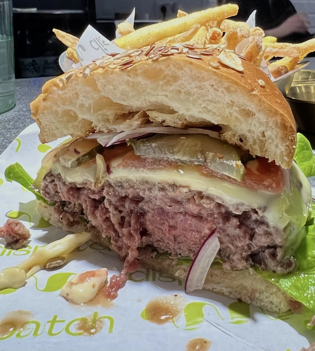 A cut-open burger with a pink beef patty, melted cheese, pickle, red onion and lettuce in a seeded bun on branded paper