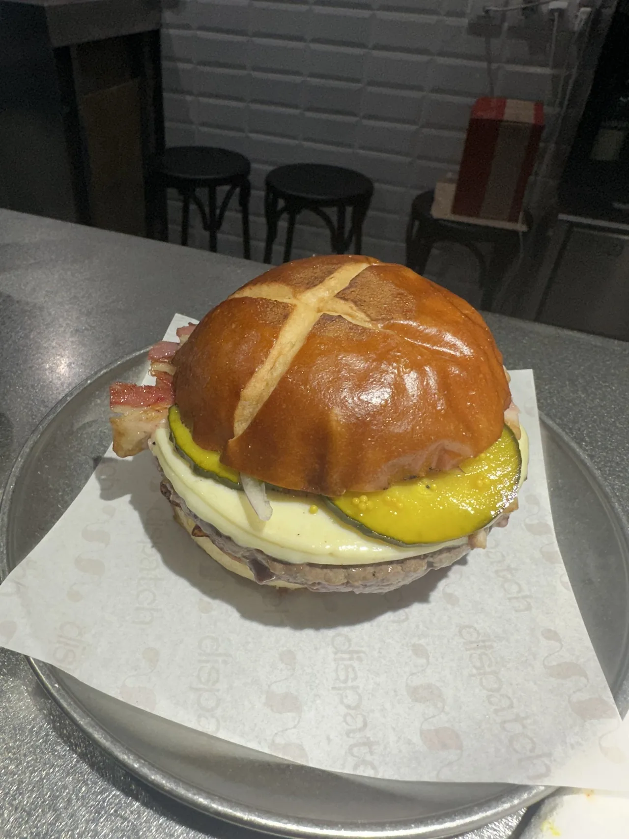 A burger with a glossy scored bun, cheese and pickle slices on a metal tray lined with paper