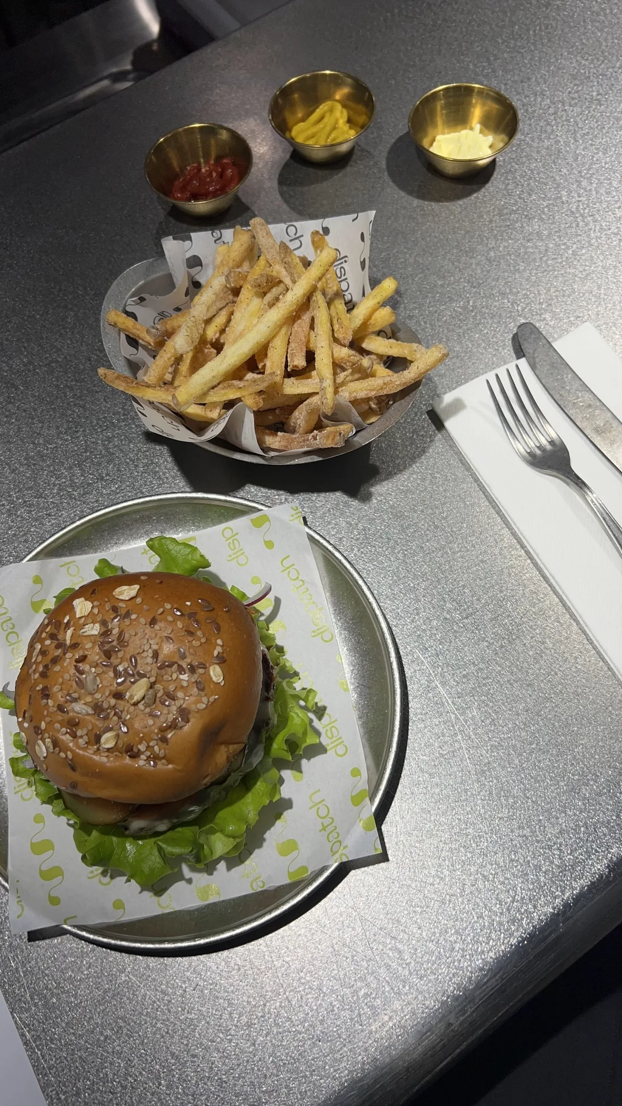 A seeded-bun burger on a lettuce leaf and a tray of fries with three sauce pots on a steel counter