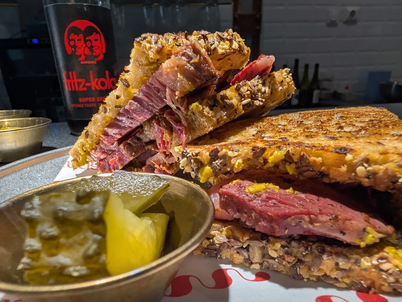 Two pastrami sandwiches on toasted bread beside a small dish of pickles