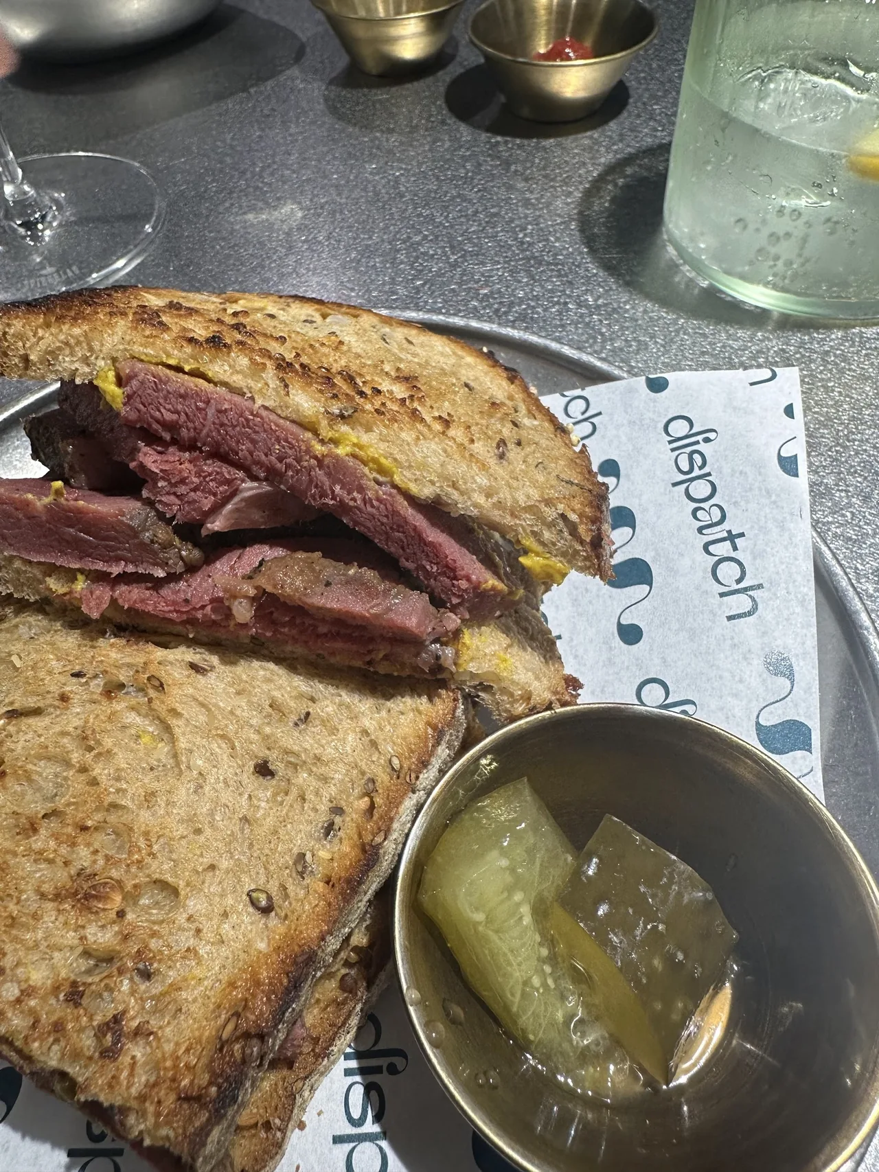 A toasted pastrami sandwich on a paper-lined plate beside a dish of pickle
