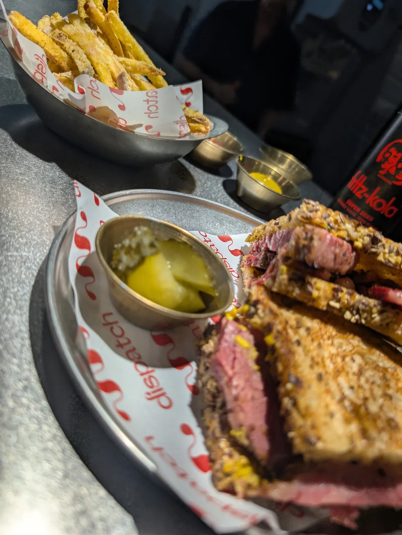 A pastrami sandwich on a paper-lined tray with a dish of pickles and fries behind