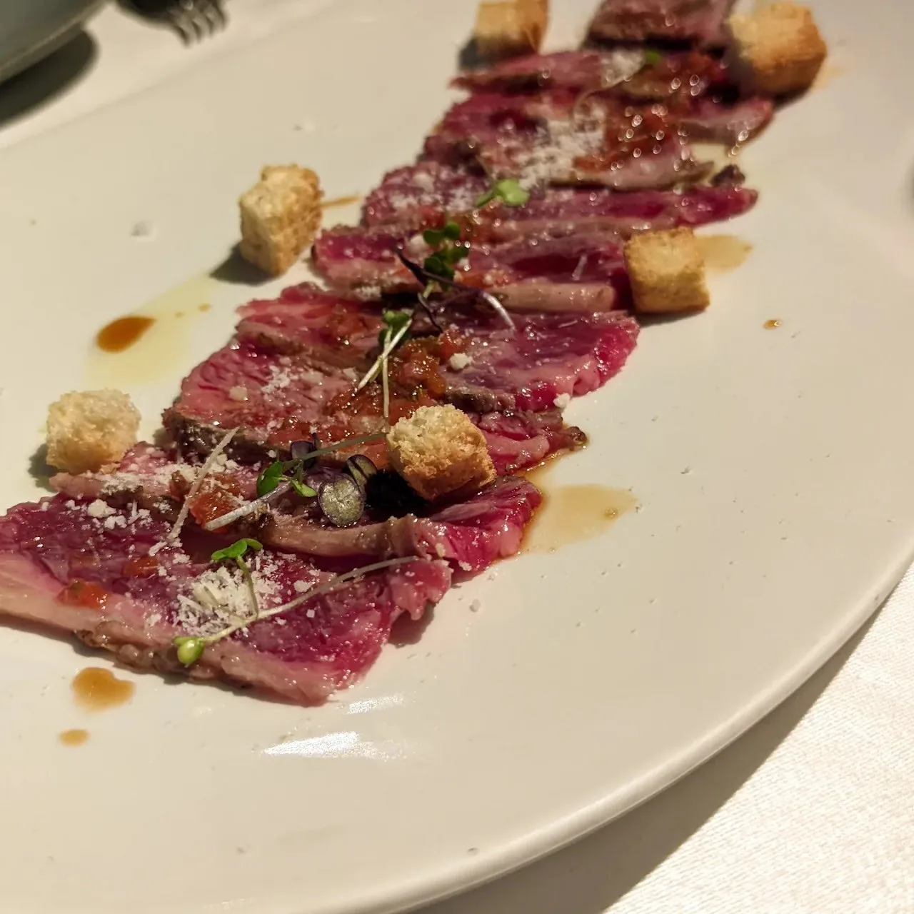 A white plate of thinly sliced rare beef with small croutons and sauce