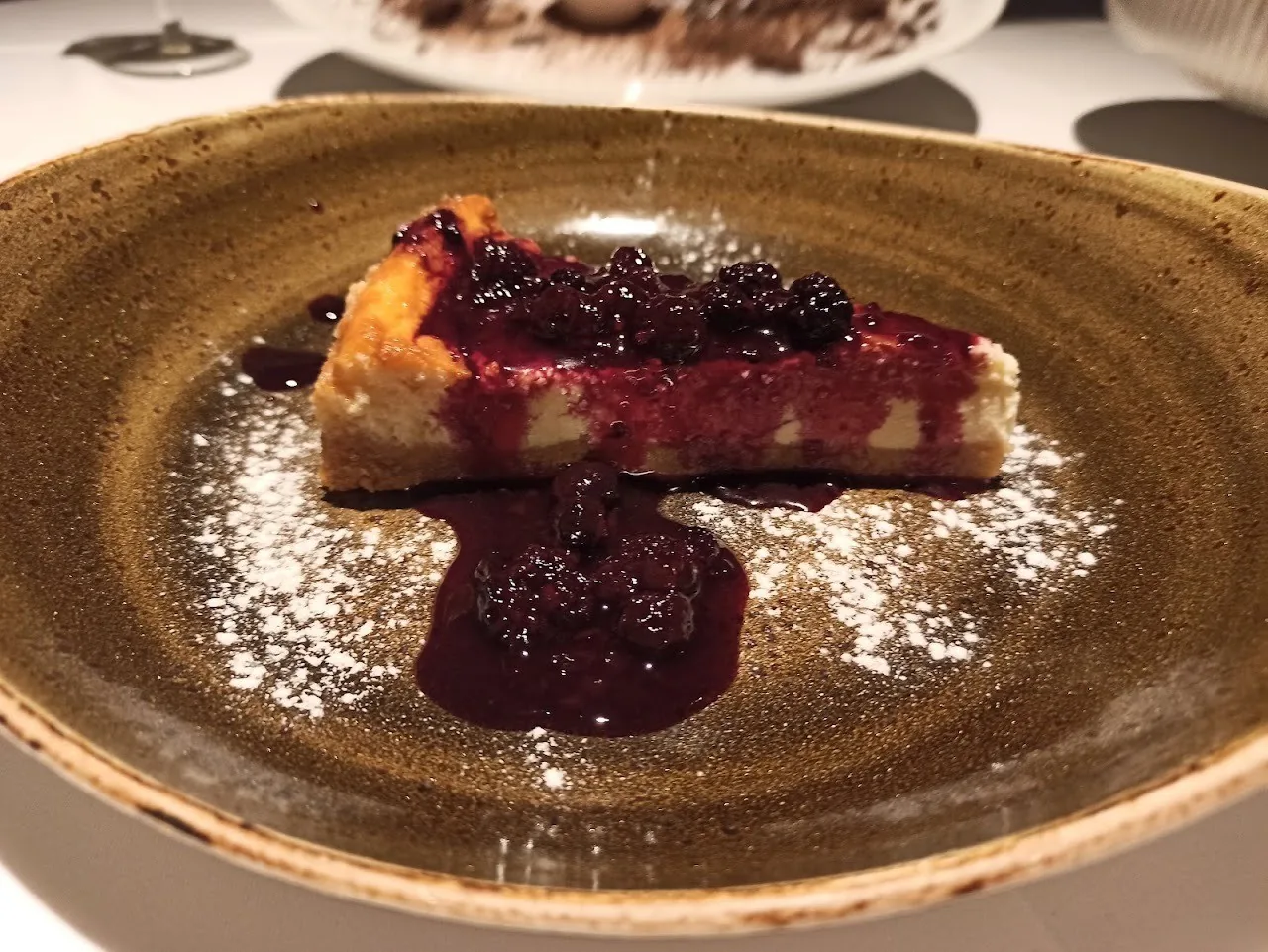 A slice of baked cheesecake with dark berry sauce on a brown plate