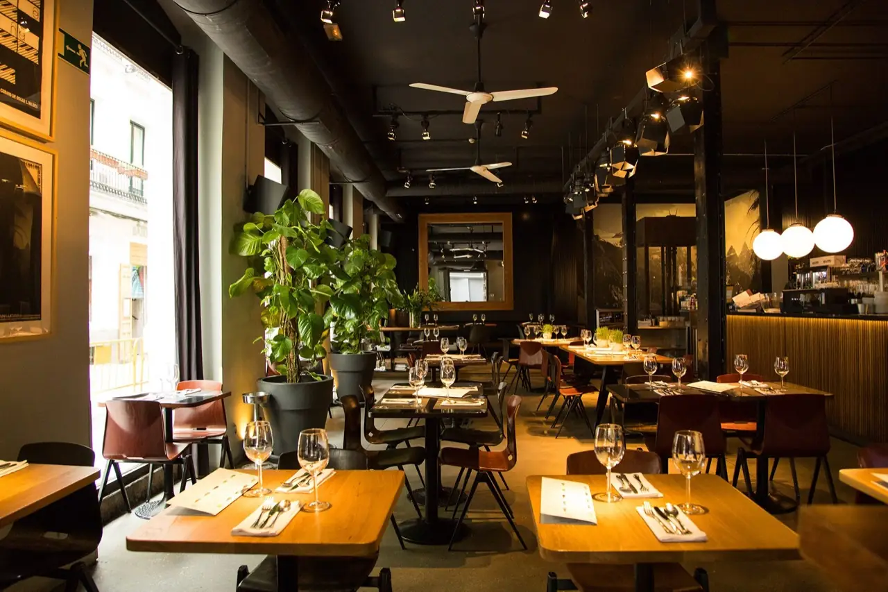 Warmly lit restaurant interior with wooden tables set with wine glasses, dark red chairs, tall potted plants by the window and a bar at the back