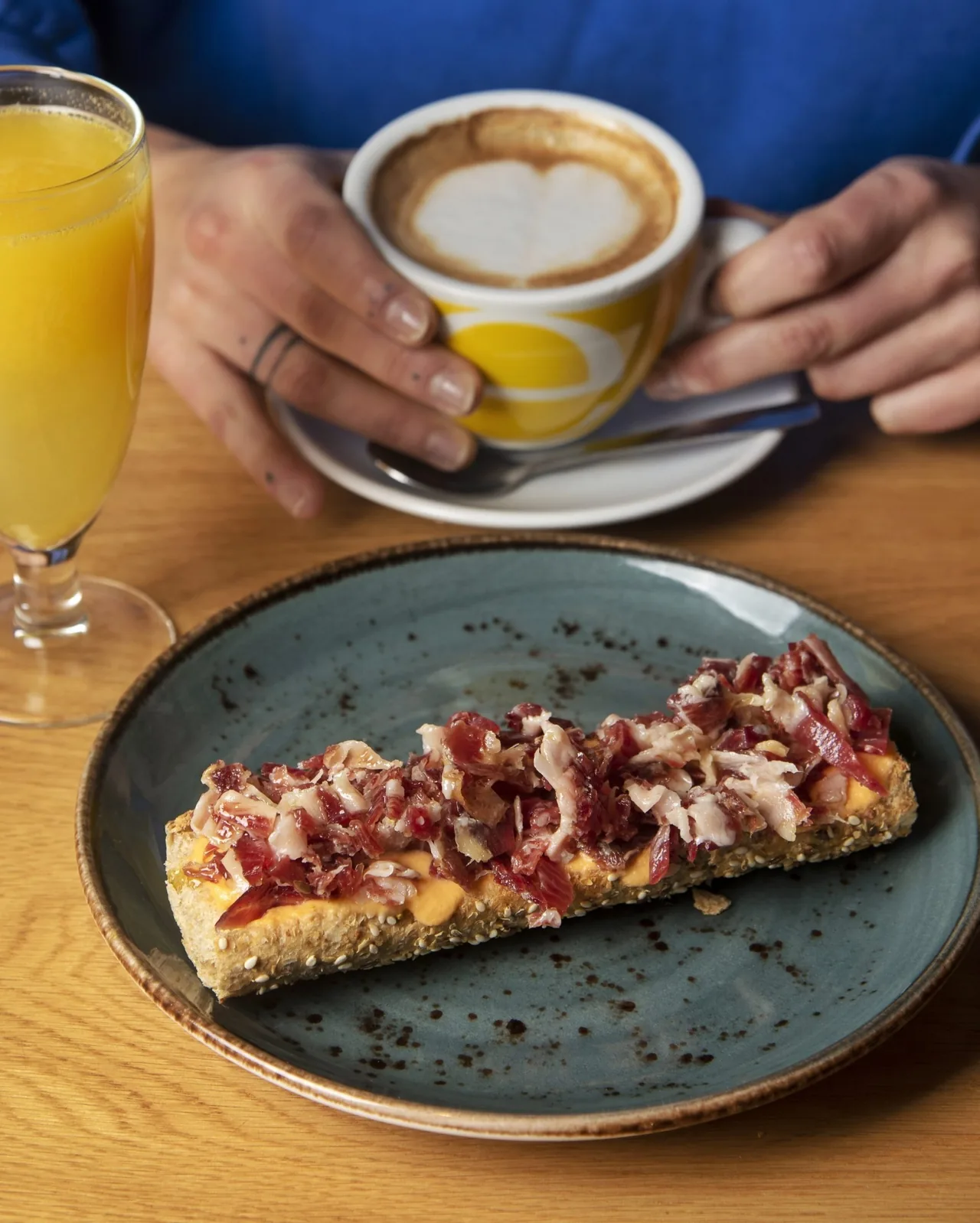 An open toast topped with slices of cured ham on a blue plate, with a cup of coffee and a glass of orange juice