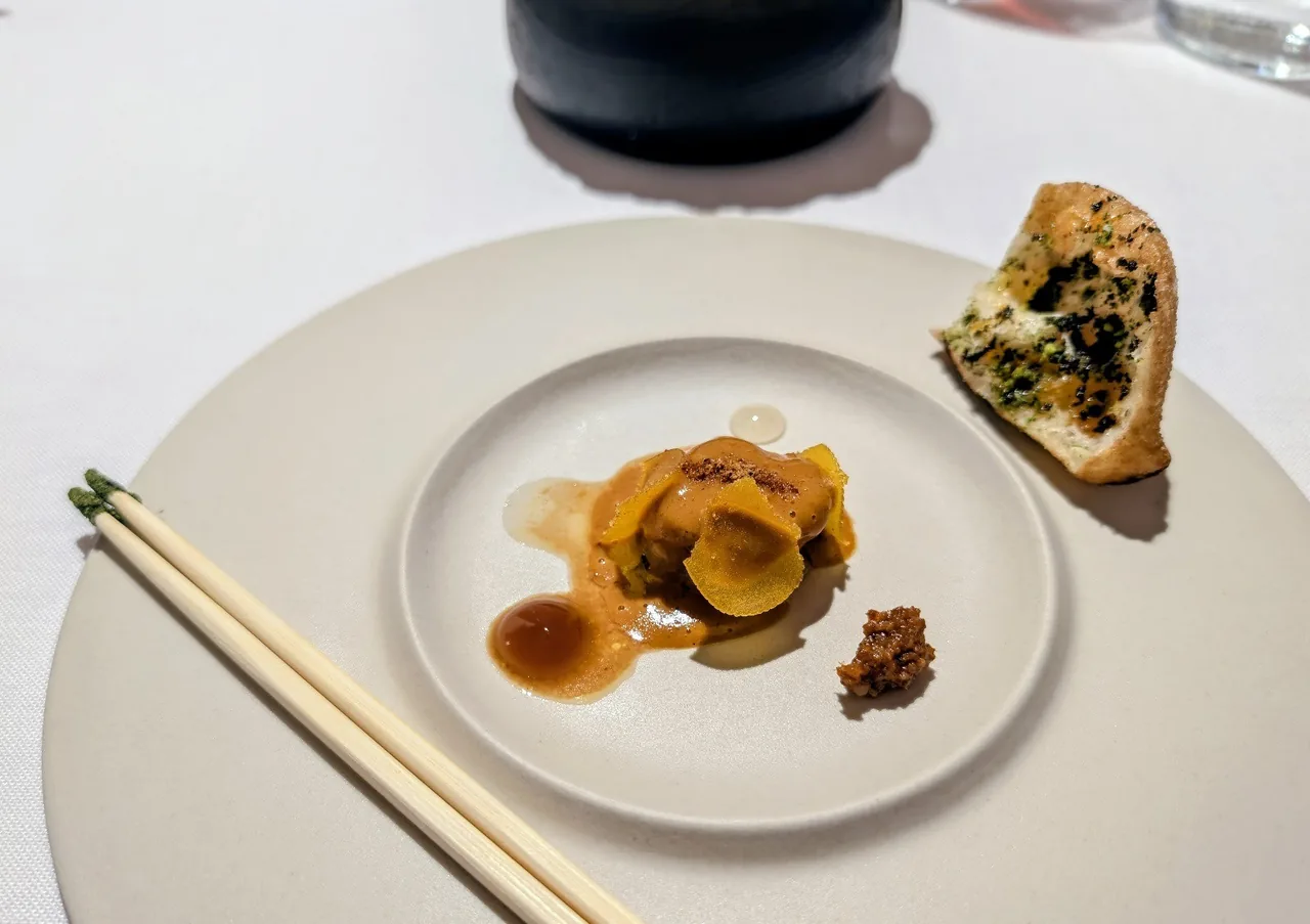 A small round plate holding a glazed dish beside a filled pastry, with chopsticks resting on a larger white plate