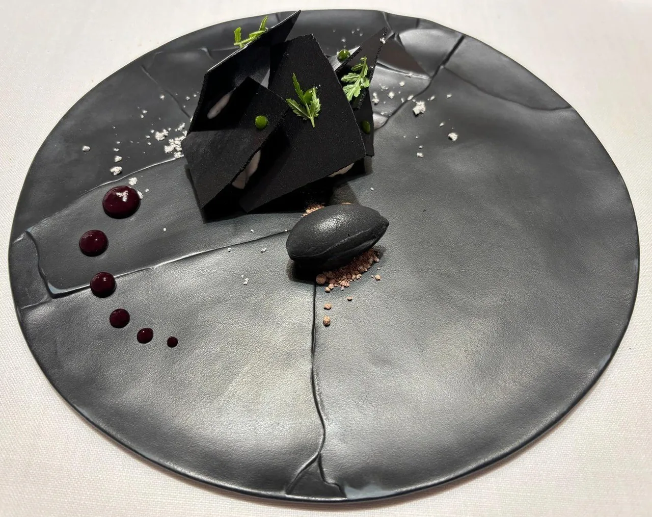 A large dark ceramic plate with a black angular element and small dots of sauce