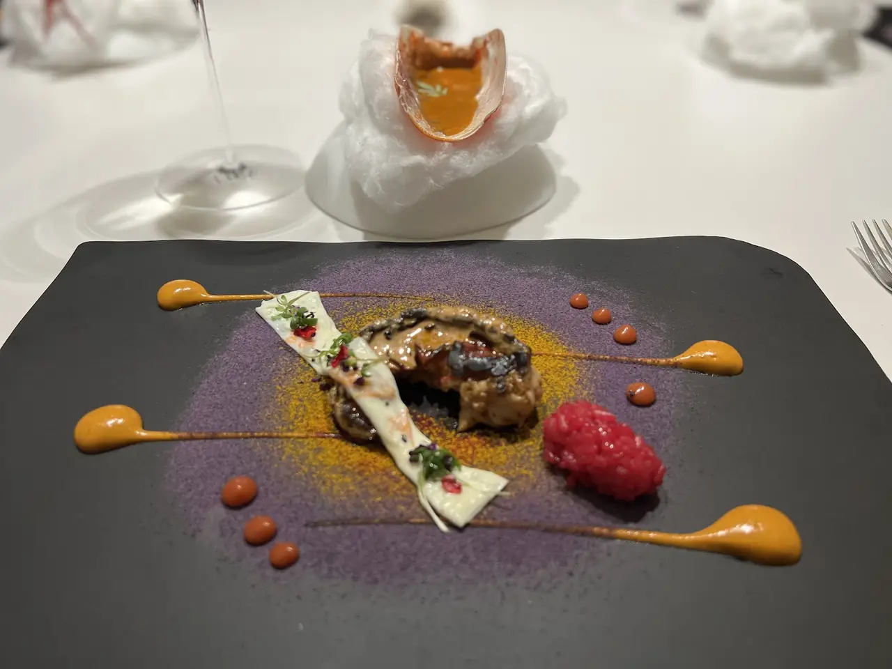 Plated dish on dark slate with sauce swirls and dots, pink puree, second course on cotton behind