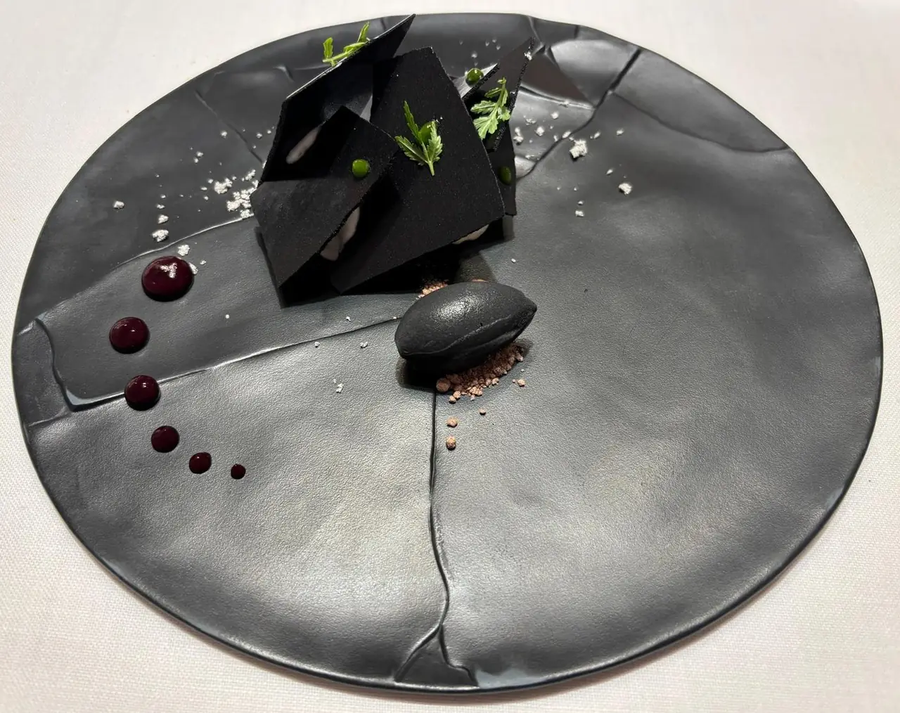 Black shard-shaped plated dish with herb garnish, dark macaron and berry sauce dots on black plate