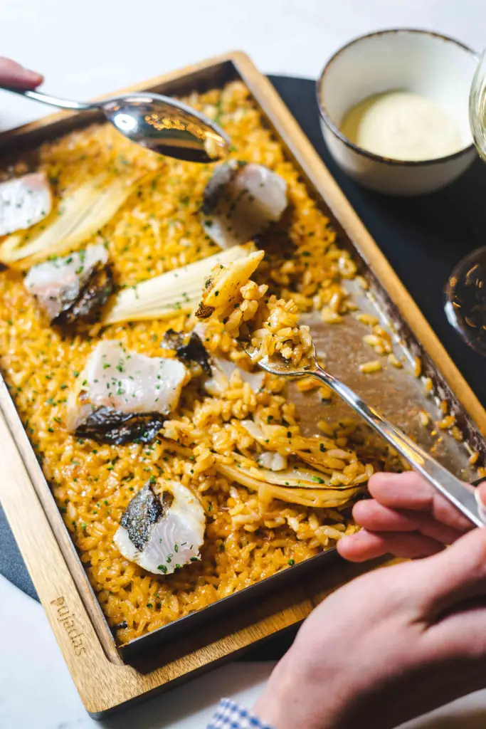 Rectangular tray of golden rice with pieces of white fish and vegetables, a forkful lifted and a bowl of aioli alongside
