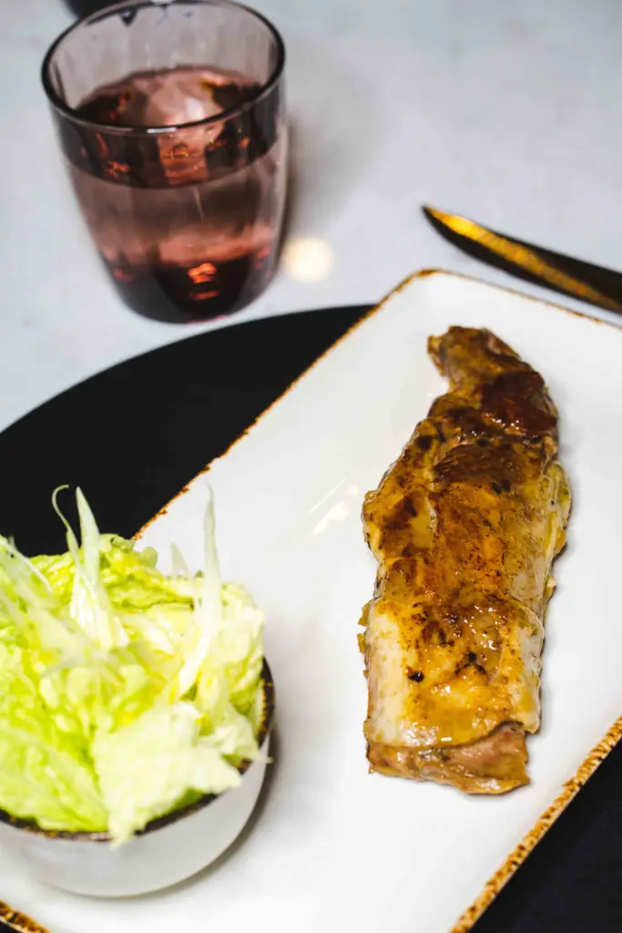 Glazed grilled fillet on a white rectangular plate with a bowl of shredded lettuce and a glass of red drink