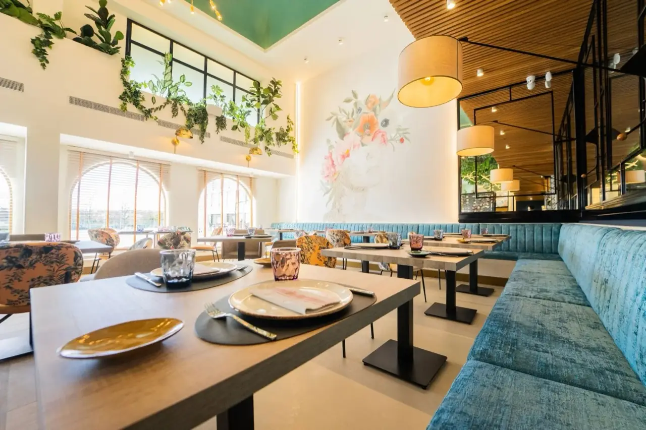 Bright dining room with set tables, teal velvet banquettes, hanging plants and a floral face mural on a white wall