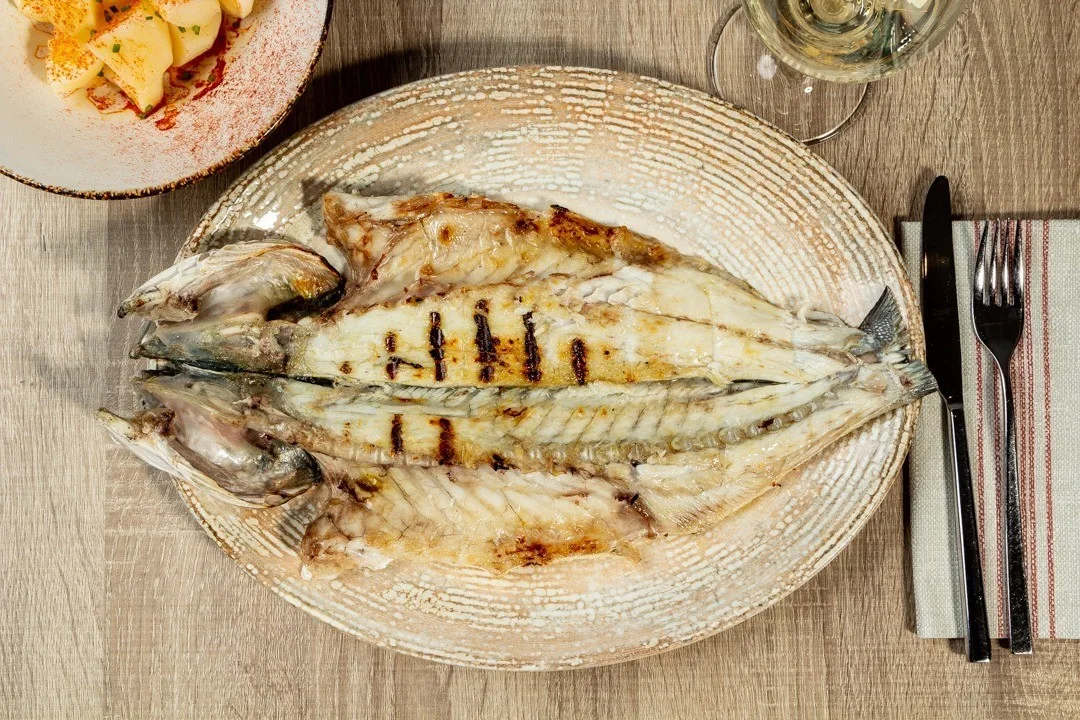 A grilled whole fish on a round mat beside cutlery