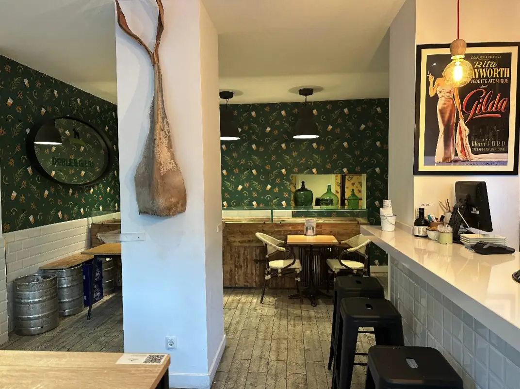 Bar interior with green botanical wallpaper, bar stools and tables, beer kegs, a Doble & Gilda mirror and a framed Rita Hayworth Gilda film poster
