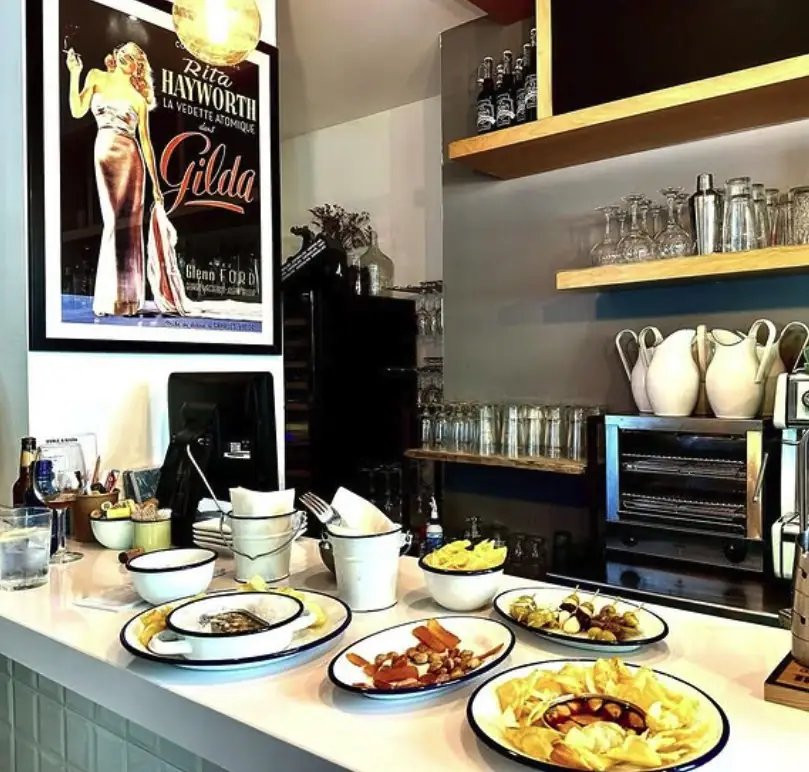 Bar counter set with several enamel plates of tapas including crisps, gildas and pintxos, under a framed Gilda film poster