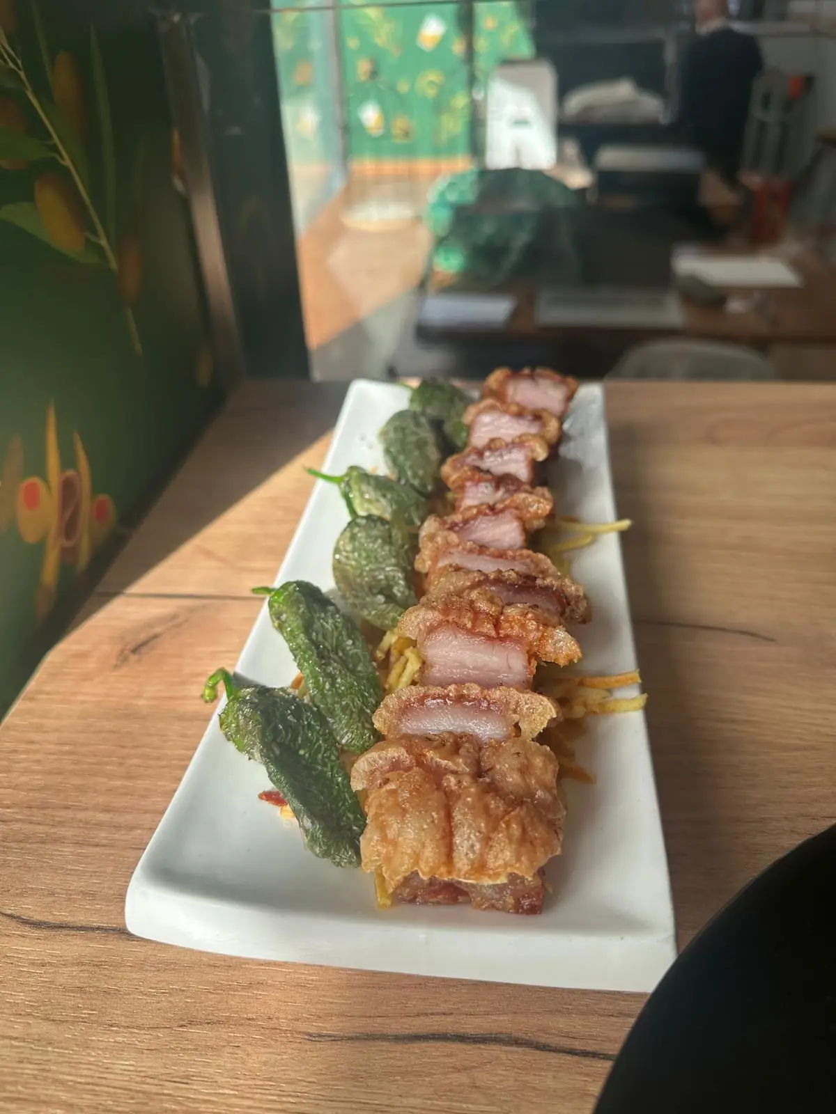 Long white plate of sliced crispy pork belly with fried padron peppers and straw potatoes on a wooden table