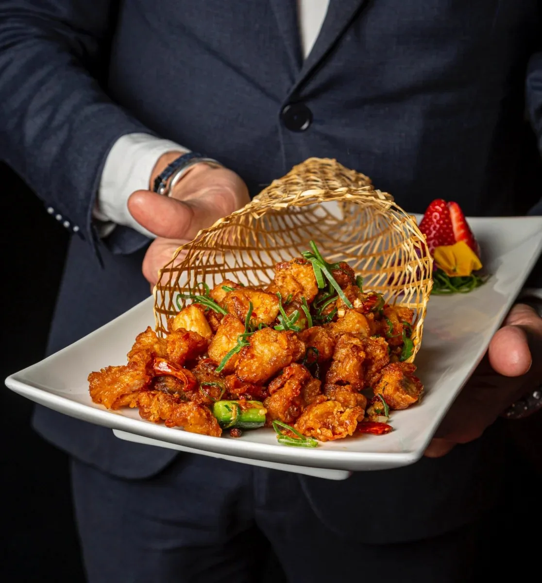 A white platter of crisp fried chicken pieces with dried chillies and sliced spring onion spilling from a woven bamboo cone, presented by a server in a navy suit