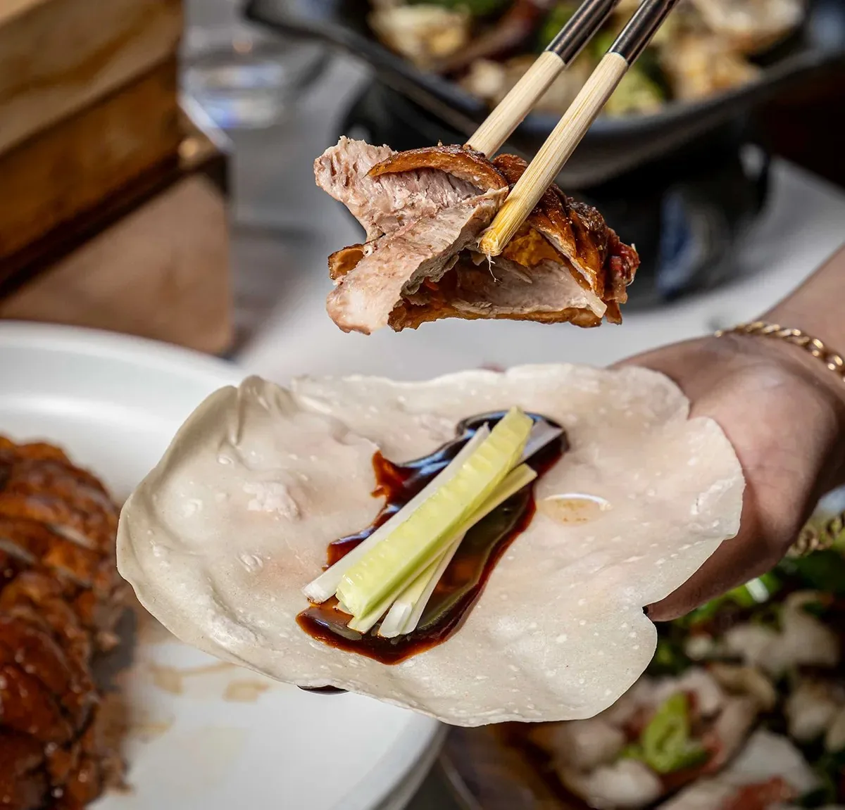 Chopsticks lifting a slice of crisp-skinned roast duck above an open pancake held in a hand, spread with dark hoisin sauce and batons of cucumber and spring onion