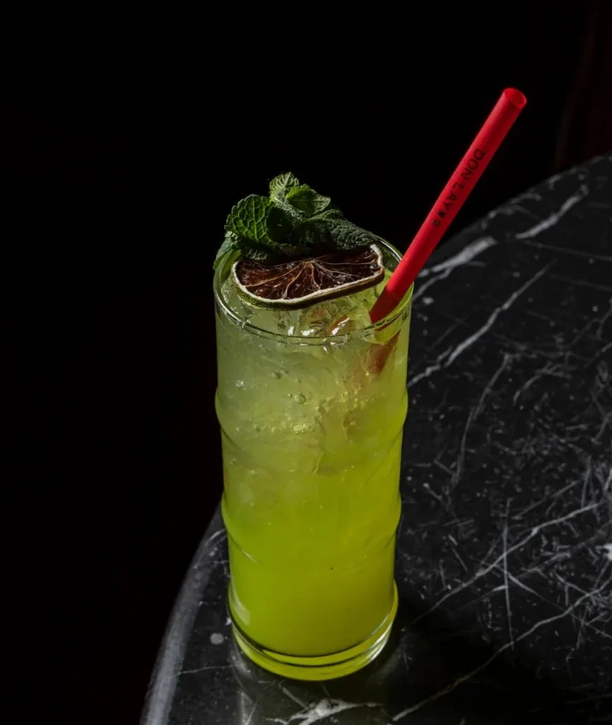 Tall glass of a bright green iced cocktail on a dark marble table, garnished with a mint sprig and a dried lime wheel, with a red straw branded Don Lay