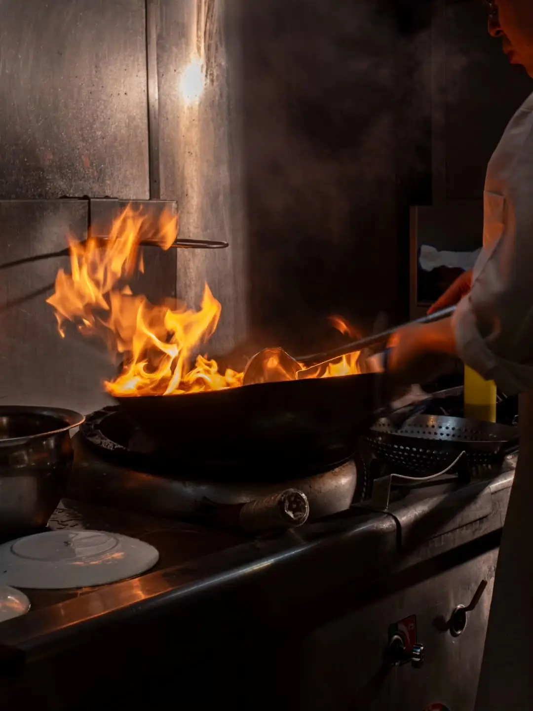 Chef tossing a flaming wok over a restaurant burner with tall orange flames