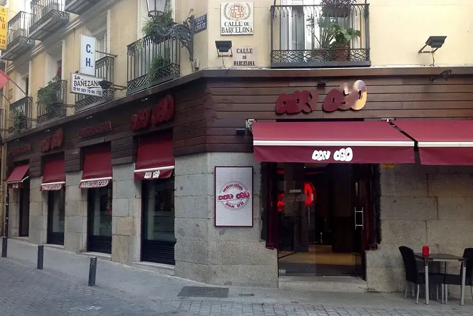 Corner exterior of Don Oso burger restaurant with maroon awnings and red signage on a granite-and-wood facade at the corner of Calle de Barcelona