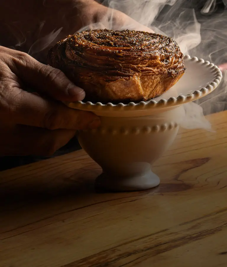 Steaming glazed swirled pastry on a white beaded pedestal held by a hand over a wooden table