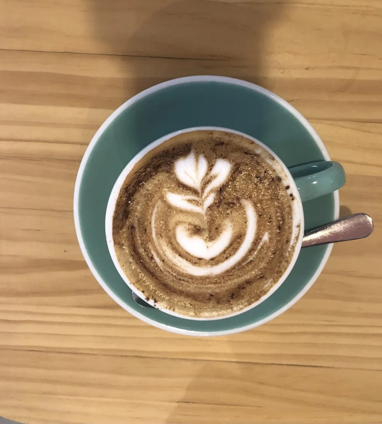 A latte with a leaf pattern in a green cup and saucer, seen from above on a wooden table
