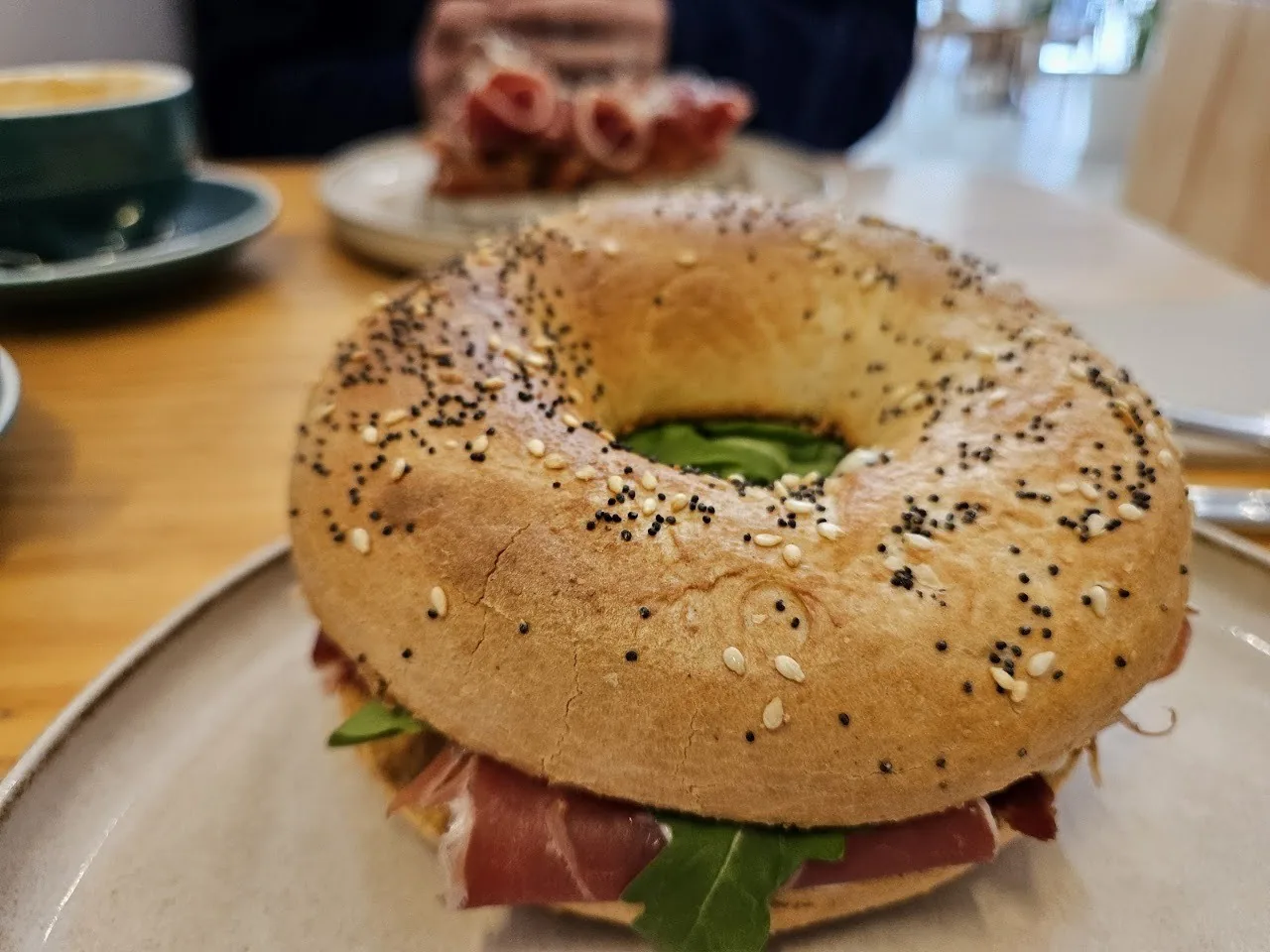 A large bagel sandwich with seeds on top, filled with cured ham and greens
