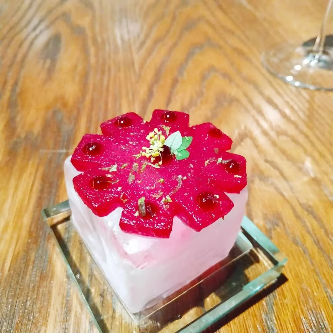 A pink glazed square dessert with red drops on a glass stand